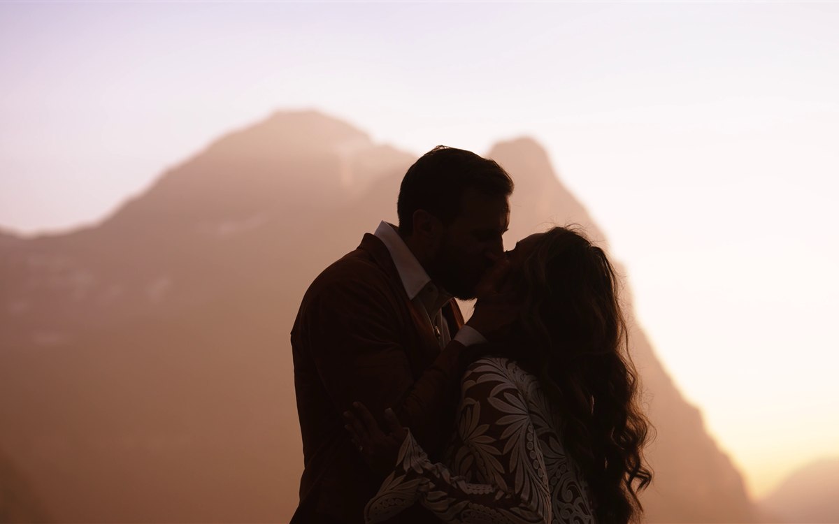 Michelle and Adam kissing at sunset with Glacier National Park's peaks silhouetted behind them